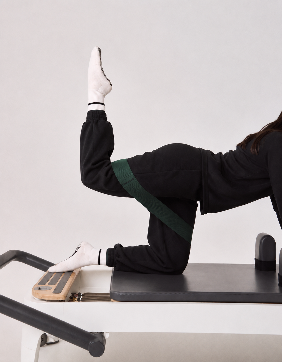 A person in a black tracksuit and white socks performs a leg exercise on a Pilates reformer machine.