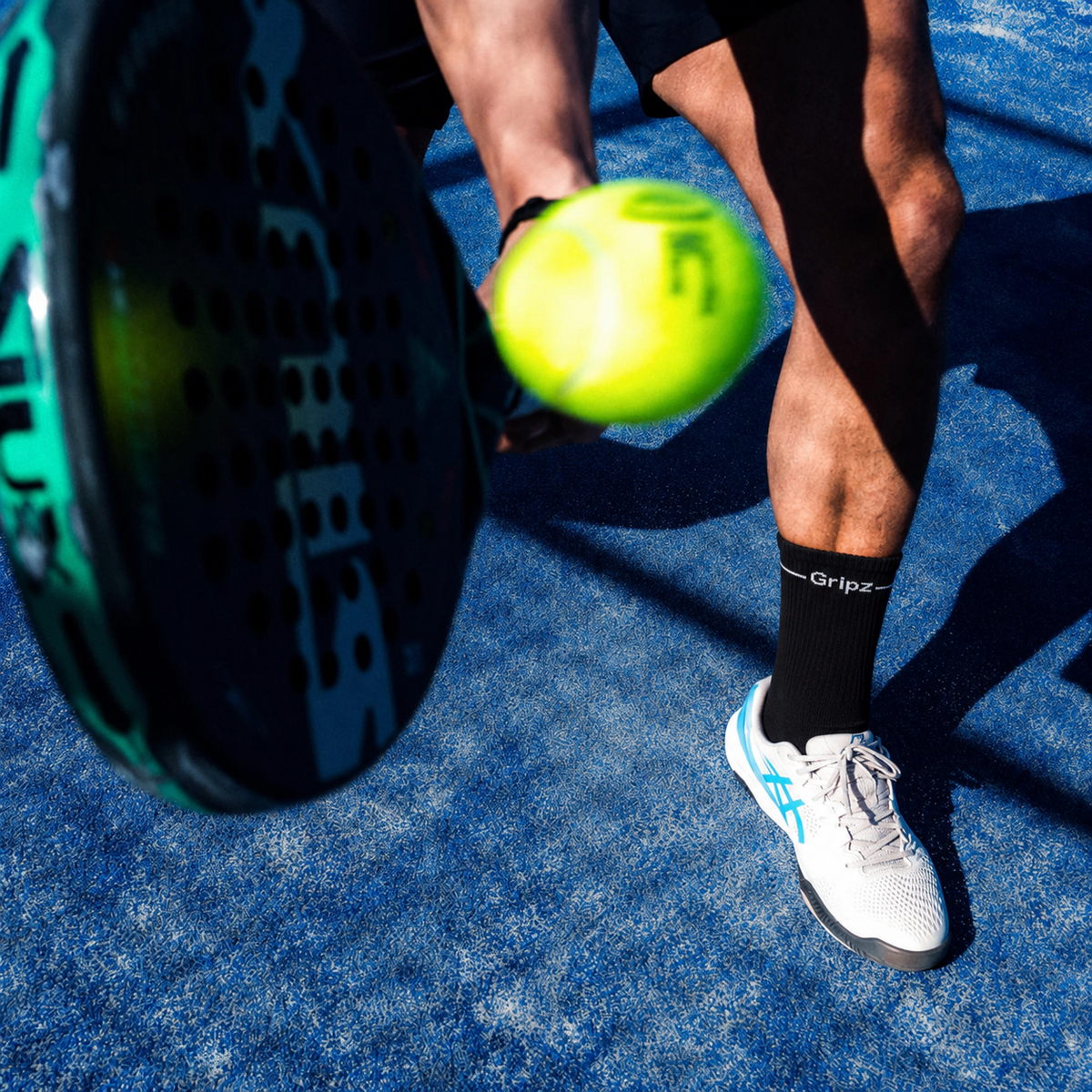 A close-up action shot of a player's leg and foot holding a bright yellow tennis ball near a paddle, on a textured blue court.