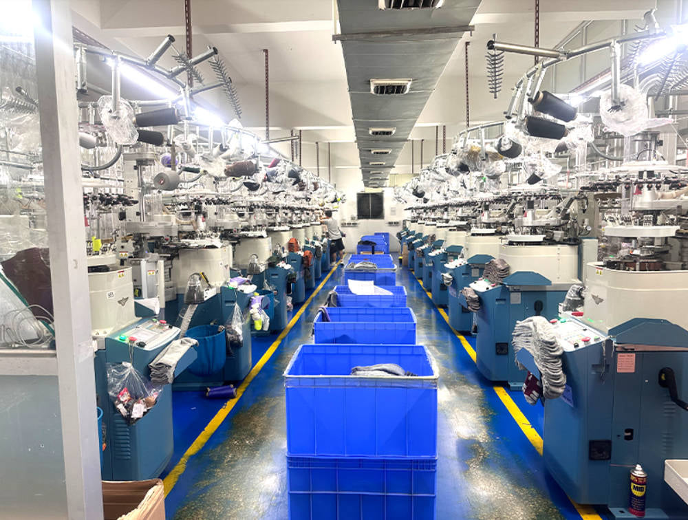 A wide view of a textile factory floor with rows of knitting machines on both sides, blue plastic bins in the center aisle, and workers visible in the distance.