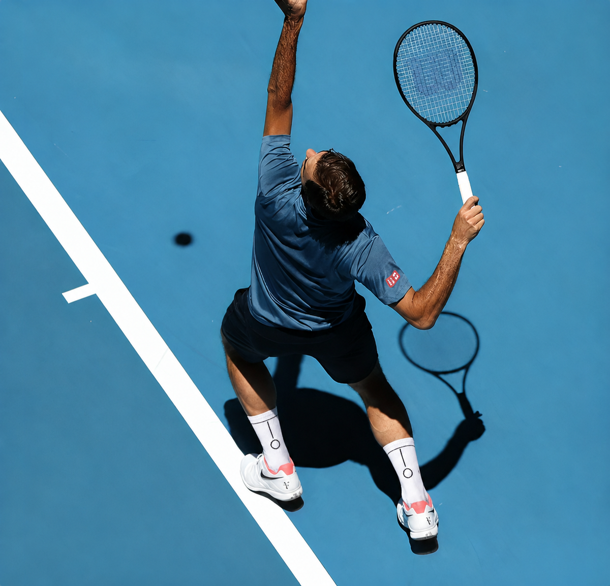 A tennis player in a blue shirt and black shorts serves a ball on a blue hard court, holding a racket.